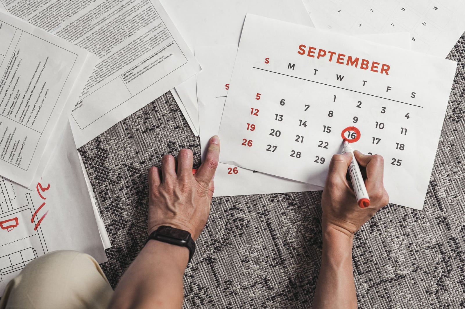 Manos sosteniendo un calendario con fechas de septiembre rodeadas, rodeado de documentos en una alfombra.