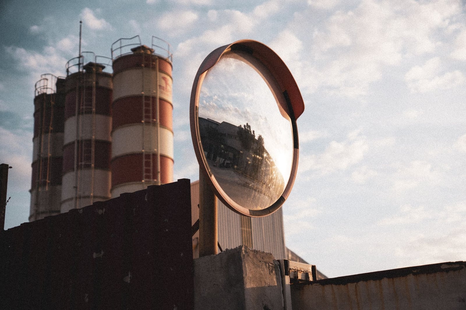 Un espejo de seguridad refleja un paisaje industrial con silos bajo un cielo nublado.